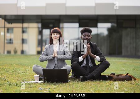 Deux jeunes gens d'affaires multiethniques en costume formel, assis sur l'herbe verte devant le bâtiment de bureau moderne et exécutant lotus pose, méditant avec les yeux fermés et le geste de nom Banque D'Images