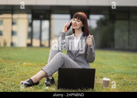 Photo d'une femme de race blanche adulte vêtue d'un vêtement d'affaires, assise sur l'herbe dans un parc urbain et parlant sur un téléphone portable tout en travaillant sur un ordinateur portable gris, en parlant et souriant et en montrant le pouce vers le haut Banque D'Images
