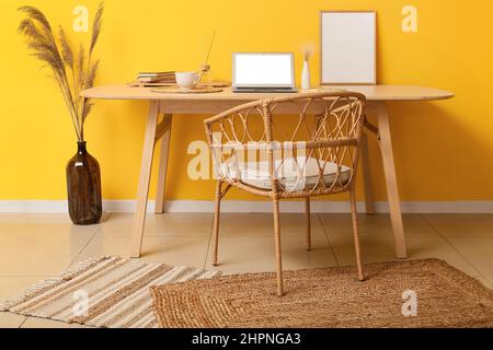 Espace de travail moderne avec ordinateur portable, tasse de café, cadre et pampas herbe dans un vase près du mur jaune Banque D'Images