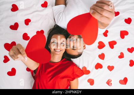 Vue de dessus d'un jeune couple heureux amoureux tenant des coeurs en papier rouge tout en étant allongé sur le lit blanc à la maison Banque D'Images