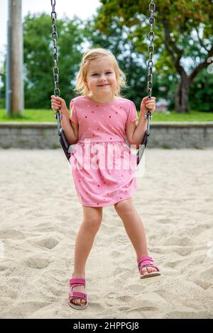 Sourire heureux petit enfant sur balançoire de chaîne. Magnifique petit enfant balançant dans l'aire de jeux dans le parc en été. Fille portant une robe rose. Banque D'Images