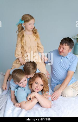 Une jeune mère heureuse se trouve sur le lit dans le chaos avec trois jeunes enfants, deux garçons sauvages et une jolie fille, et leur père est assis à côté l'un de l'autre.Un week-end amusant avec votre famille. Banque D'Images