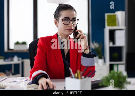 Une femme d'affaires confiante est assise sur un téléphone intelligent au bureau. Manager avec des lunettes travaillant dans le bureau de démarrage tout en parlant au téléphone. Employé en blouson rouge lors d'une conversation sur téléphone portable. Banque D'Images