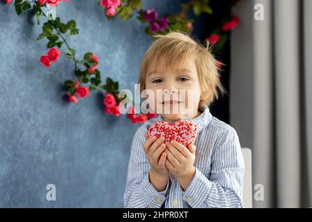 Mignon petit enfant blond, petit garçon d'âge préscolaire, mangeant le donut rose en forme de coeur, fait pour la Saint-Valentin, fond de Saint-Valentin Banque D'Images