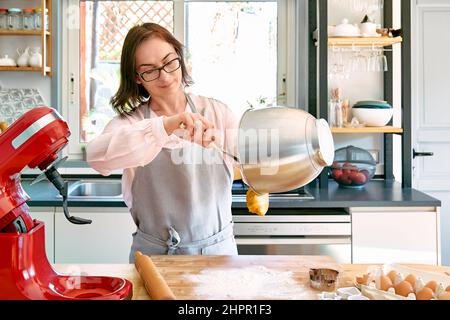 Femme portant un tablier de cuisson des biscuits dans une cuisine confortable. La femme de ménage prend la pâte du bol du batteur planétaire. Gâteaux ou tartes faits maison. Banque D'Images