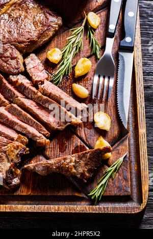 steaks de bœuf de bœuf juteux poêlé, un tout et un coupé en tranches avec gousses d'ail frites et romarin sur panneau de bois avec sauce barbecue, vie verticale Banque D'Images