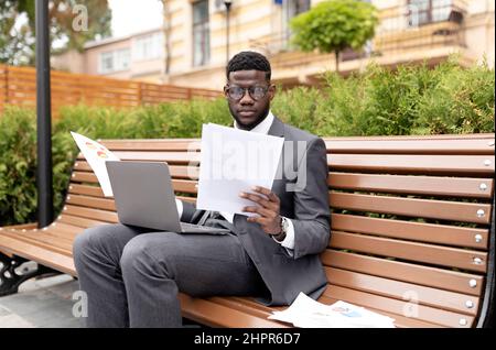 Concept de carrière professionnelle. Un entrepreneur américain sérieux travaillant sur ordinateur portable et lisant des documents, assis sur un banc Banque D'Images