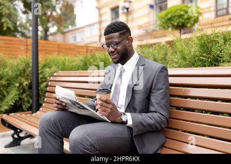 Un homme d'affaires afro-américain confiant lisant les actualités du jour dans les journaux et buvant du café à emporter, assis sur son banc Banque D'Images