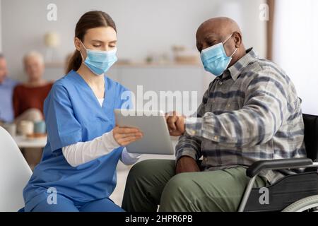 Femme en vêtements de travail aidant homme noir senior à utiliser une tablette numérique Banque D'Images