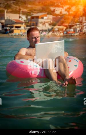 Travailler toujours et partout. Portrait humoristique d'un indépendant travaillant avec un ordinateur portable à l'extérieur dans l'eau de la mer tropicale. Banque D'Images