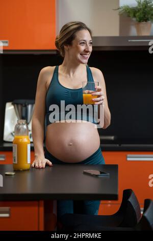 Femme blonde enceinte buvant un jus d'orange dans la cuisine Banque D'Images