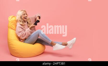 Joyeuse femme jouant des jeux vidéo avec joystick, assis sur un fauteuil de beanbag sur fond rose studio, espace libre Banque D'Images