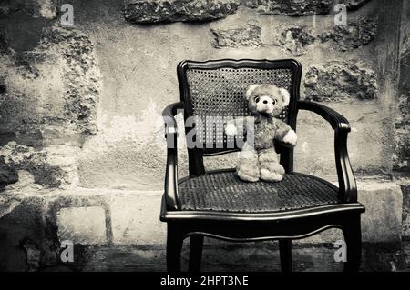 Ours en peluche assis sur une chaise et mur en pierre rugueuse à l'arrière-plan. Photo conceptuelle historique noir blanc. Banque D'Images