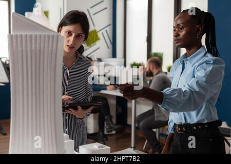 Femme architecte afro-américaine pointant un crayon sur le modèle architectural en mousse de gratte-ciel, debout à côté de l'ingénieur de collègue tenant une tablette dans un bureau moderne. Architectes travaillant en équipe. Banque D'Images