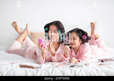 Bonne jeune femme japonaise et adolescente en pyjama et les curlers se trouvent avec des cosmétiques sur le lit, s'amuser Banque D'Images