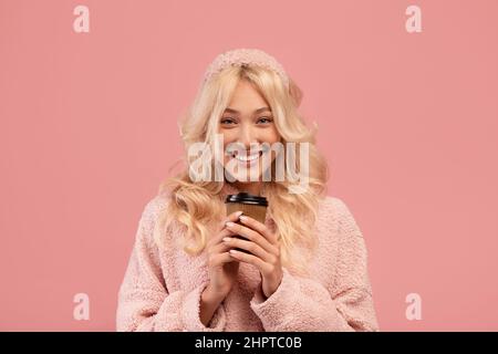 Boisson chaude préférée et pause. Bonne dame tenant une tasse en papier à emporter et souriant à l'appareil photo, debout sur fond rose Banque D'Images