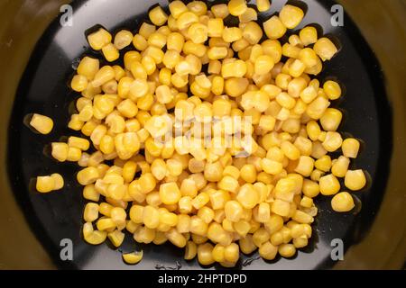 Grains doux jaune vif de maïs en conserve dans une plaque en céramique noire, macro, vue de dessus. Banque D'Images