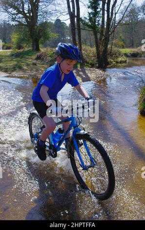 Un garçon fait du vélo en toute confiance à travers le Fletchers Water Ford dans le parc national de New Forest, avec plaisir et éclaboussures. Banque D'Images