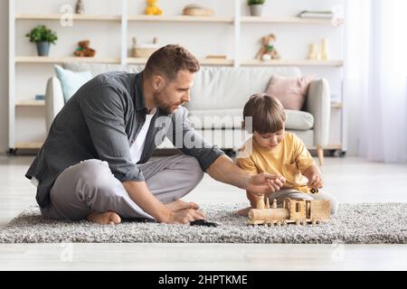 Un jeune père heureux passe du temps avec son petit fils mignon à la maison, assis sur le sol et jouant du train en bois, espace libre Banque D'Images