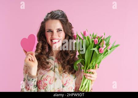 femme élégante et souriante d'âge moyen avec de longs cheveux de brunette ondulés avec bouquet de tulipes et coeur rose isolé sur rose. Banque D'Images