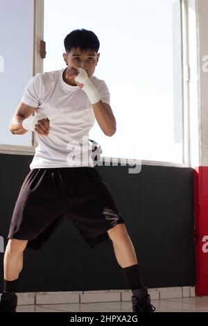 un jeune boxeur portant un t-shirt blanc à l'ombre de boxe avec ses poings bandés pendant son entraînement à l'intérieur de la salle de boxe. Banque D'Images