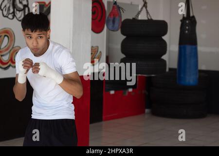 un jeune boxeur portant un t-shirt blanc à l'ombre de boxe avec ses poings bandés pendant son entraînement à l'intérieur de la salle de boxe. Banque D'Images