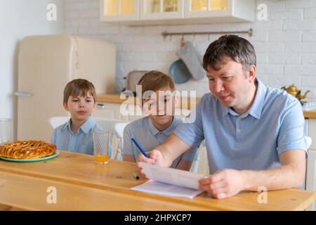 Un père caucasien attentionné assis à table avec ses fils et aidant à faire ses devoirs à la maison, un père européen aimant et un petit étudiant ensemble dans la cuisine en quarantaine, concept de scolarisation à domicile Banque D'Images