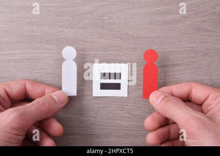 Égalité entre la diversité raciale et les mains tenant deux découpes de papier en forme de personne et une affiche avec signe égal sur une table en bois. Vue du dessus. Banque D'Images