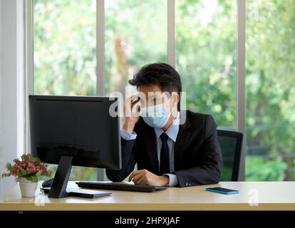 De jeunes hommes d'affaires asiatiques avec masque de visage touchent son temple d'une main tout en travaillant dans le bureau. Banque D'Images
