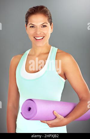 Il est agréable d'être en forme. Portrait d'une jeune femme tenant un tapis de yoga sur fond gris. Banque D'Images