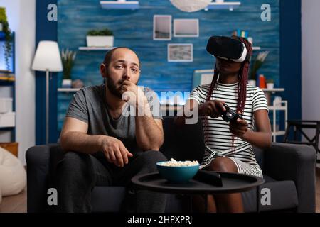 Femme afro-américaine appréciant la réalité virtuelle tandis que l'homme caucasien regardant et de manger des collations. Jeune amie jouant à des jeux VR en utilisant un casque de haute technologie pour la réalité simulée Banque D'Images