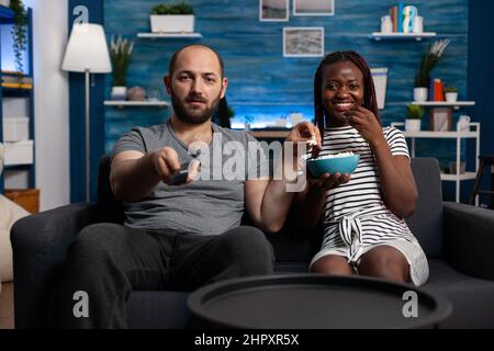 Un jeune couple multiethnique regardant un spectacle télévisé tout en partageant un bol de pop-corn. Homme caucasien changer de chaîne de télévision à l'aide de la télécommande tandis que femme afro-américaine offre des collations. Banque D'Images