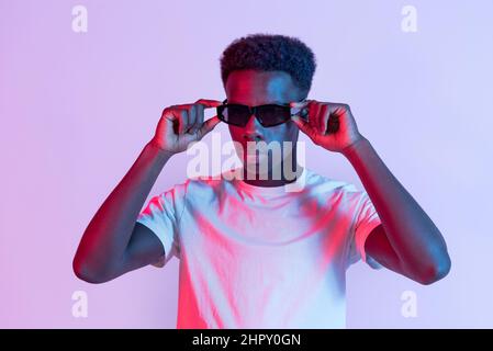 Modèle africain américain sérieux en tant que t-shirt debout et réglage de lunettes de soleil noires en studio avec éclairage coloré Banque D'Images
