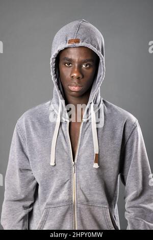 Homme afro-américain sérieux à capuche debout et regardant l'appareil photo sur fond gris en studio Banque D'Images