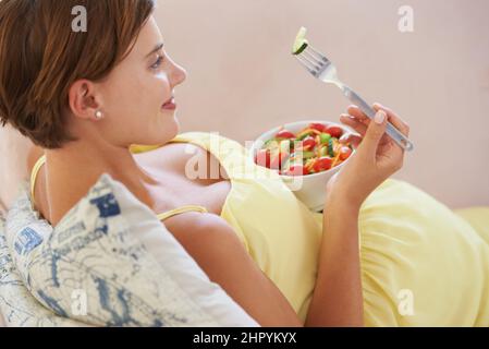 Savoureux. Une jeune femme enceinte appréciant une salade saine tout en se relaxant sur le canapé. Banque D'Images