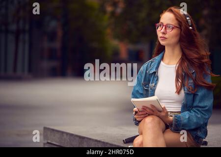 Une femme vlogger ou un social influenceur portant des écouteurs voyageant dans la ville faisant des notes dans le livre Banque D'Images