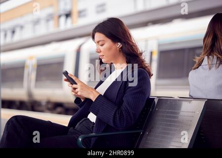 Deux femmes d'affaires qui se font du travail attendent le train sur la plate-forme de la gare qui regarde les téléphones mobiles Banque D'Images