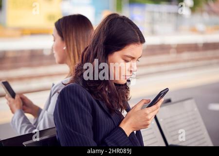 Deux femmes d'affaires qui se font du travail attendent le train sur la plate-forme de la gare qui regarde les téléphones mobiles Banque D'Images