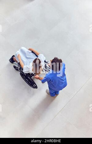 Un coup de tête d'une infirmière portant des produits de nettoyage poussant une patiente en fauteuil roulant dans le bâtiment de l'hôpital Banque D'Images
