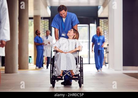 Infirmière masculine portant des produits de nettoyage poussant la patiente en fauteuil roulant dans le bâtiment de l'hôpital Banque D'Images