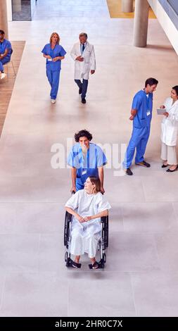 Un coup de tête d'une infirmière portant des produits de nettoyage poussant une patiente en fauteuil roulant dans le bâtiment de l'hôpital Banque D'Images