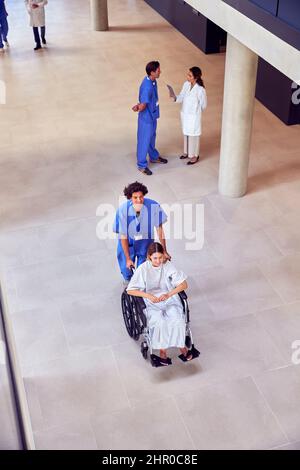 Un coup de tête d'une infirmière portant des produits de nettoyage poussant une patiente en fauteuil roulant dans le bâtiment de l'hôpital Banque D'Images
