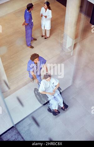 Un coup de tête d'une infirmière portant des produits de nettoyage poussant une patiente en fauteuil roulant dans le bâtiment de l'hôpital Banque D'Images