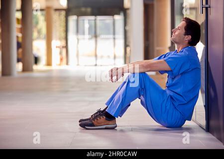 Travailleur médical fatigué et surtravaillé dans des exfoliants assis sur le plancher du couloir de l'hôpital pendant la pause Banque D'Images