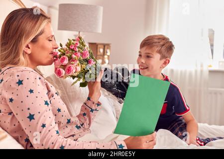 Fils aimant mère surprenante au lit avec fleurs et carte maison à l'anniversaire ou à la fête des mères Banque D'Images