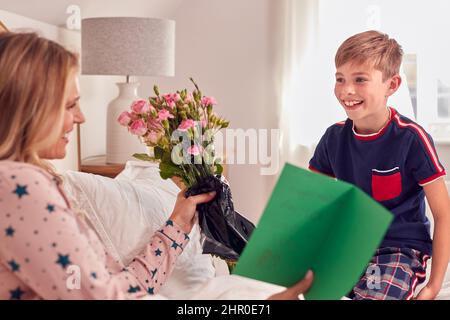 Fils aimant mère surprenante au lit avec fleurs et carte maison à l'anniversaire ou à la fête des mères Banque D'Images