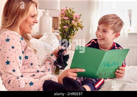 Fils aimant mère surprenante au lit avec fleurs et carte maison à l'anniversaire ou à la fête des mères Banque D'Images