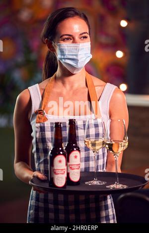 Portrait d'un serveur féminin portant un masque facial tenant un plateau de boissons au Cool Bar ou au Club Banque D'Images