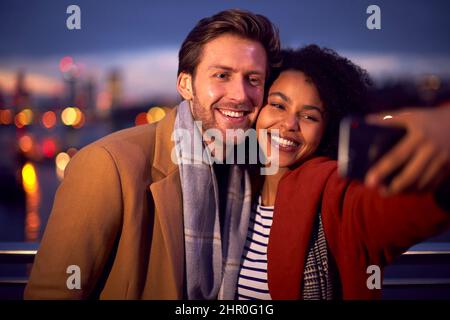 Couple contre les lumières de la ville en plein air portant des manteaux et des Scarves posant pour Selfie au téléphone à Dusk Banque D'Images