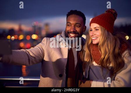 Couple contre les lumières de la ville en plein air portant des manteaux et des Scarves posant pour Selfie au téléphone à Dusk Banque D'Images